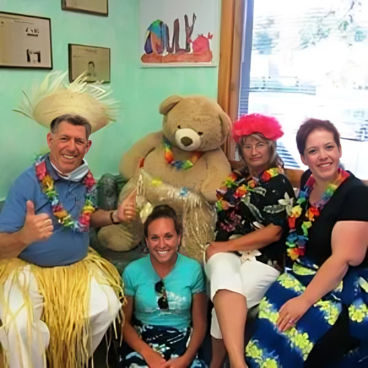 Dental office staff in colorful costumes with giant teddy bear, celebrating with child patient in clinic room.