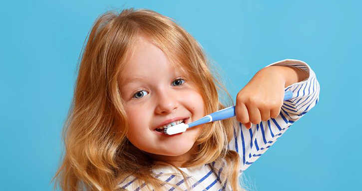 Young child with red hair brushing teeth against blue background, smiling brightly.