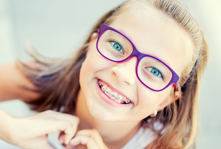 Young girl wearing purple glasses smiling brightly at camera, displaying healthy teeth.