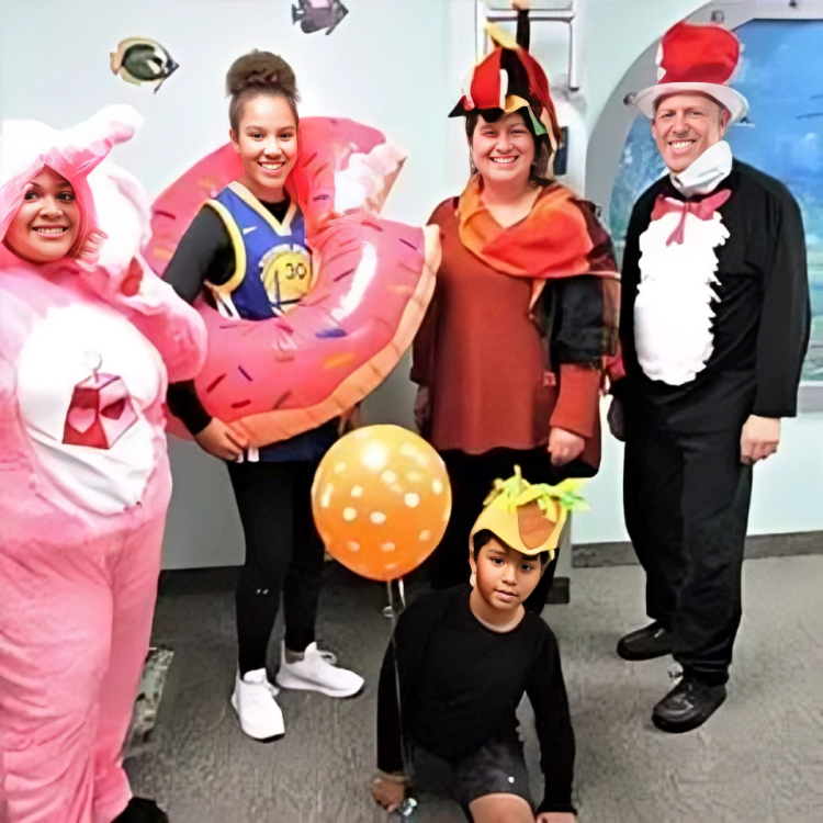 Group of children and adults in colorful Halloween costumes celebrating in an indoor office setting.