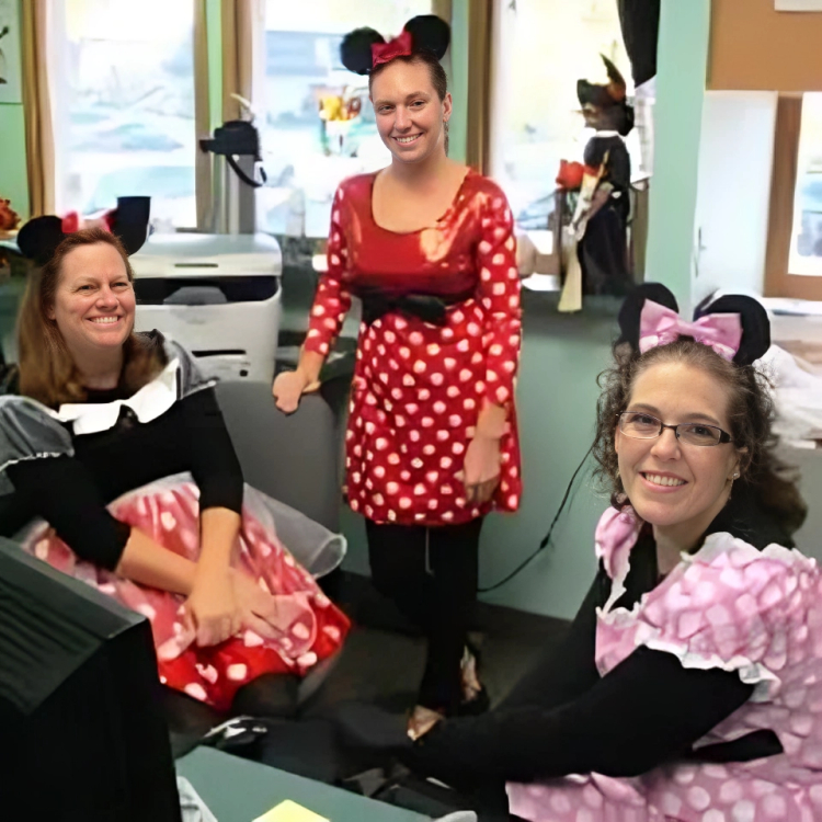 Three people wearing Minnie Mouse ears and red polka dot outfits smile in a dental office classroom setting.