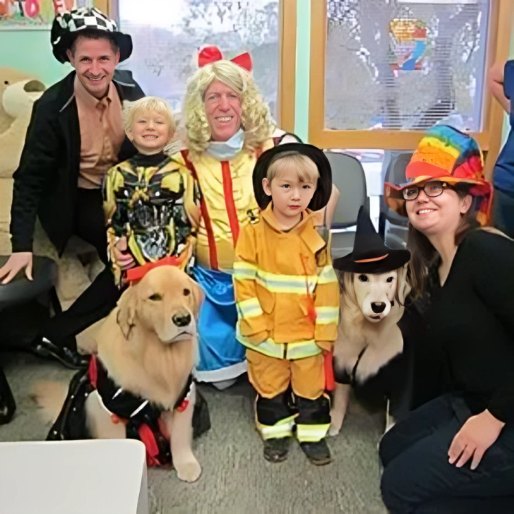 Adults and children in Halloween costumes with two dogs indoors at dental office celebration.