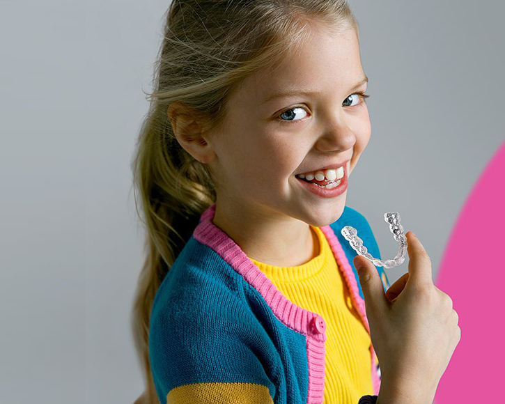 Young girl smiling brightly while holding orthodontic braces, wearing colorful clothing against neutral background.