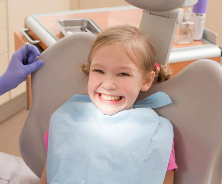 Young child smiling in dental chair wearing bib during pediatric dental examination.
