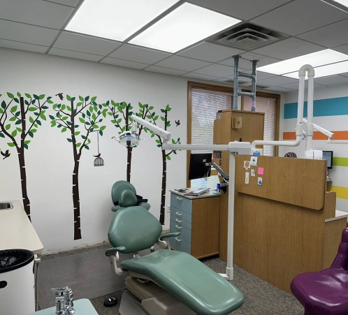 Modern pediatric dental office with treatment chair, green topiary wall mural, and wooden cabinetry.